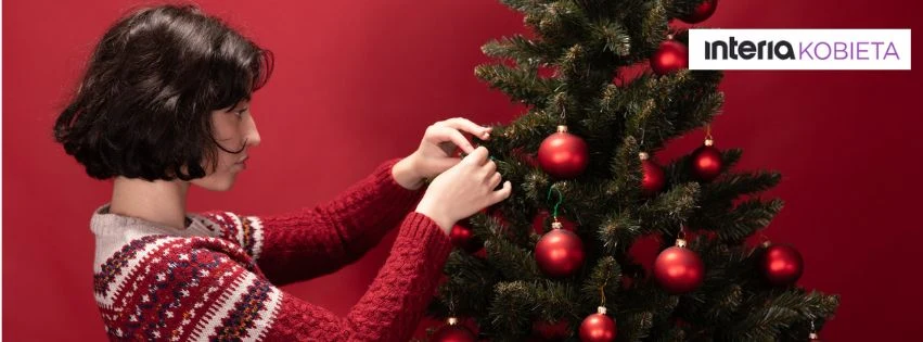 Sięgnij po inspiracje, które pomogą stworzyć ciepłą, świąteczną atmosferę Kobieta w czerwonym swetrze dekoruje choinkę czerwonymi bombkami na tle czerwonej ściany.
