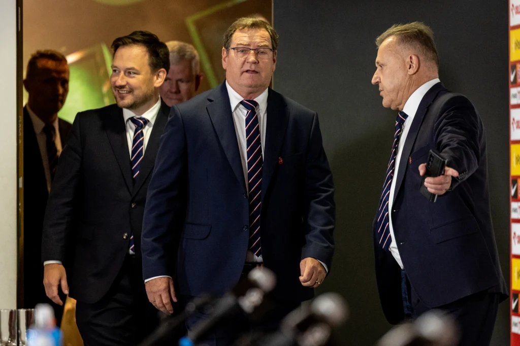 Trzech mężczyzn w garniturach stoi razem w dobrze oświetlonym pomieszczeniu. Dwóch z nich uśmiecha się, jeden wskazuje dłonią kierunek, a drugi spogląda przed siebie z neutralnym wyrazem twarzy. W tle widoczne są zamazane sylwetki innych osób oraz tabl...