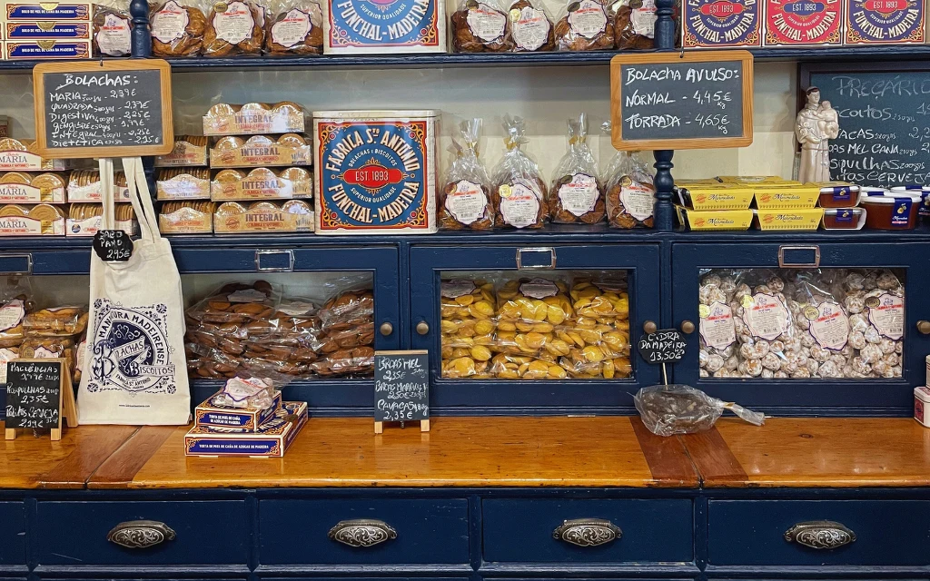 Półki z tradycyjnymi portugalskimi wypiekami i słodkościami zapakowanymi w przezroczyste torby i estetyczne pudełka, oddzielone tabliczkami z cenami, na drewnianym blacie widoczne dodatkowe paczki i torba z nadrukiem.