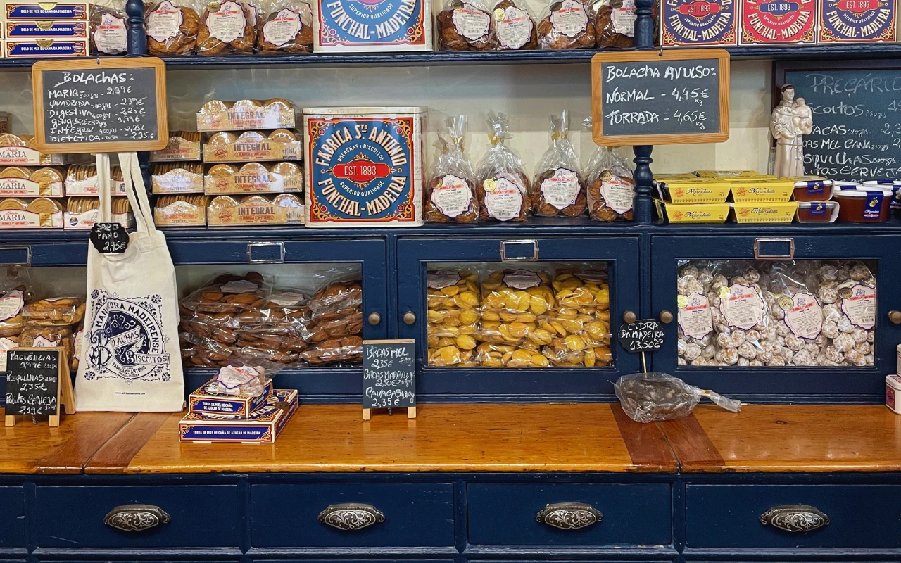 Bolo de mel można kupić w maderskich cukierniach i piekarniach Półki z tradycyjnymi portugalskimi wypiekami i słodkościami zapakowanymi w przezroczyste torby i estetyczne pudełka, oddzielone tabliczkami z cenami, na drewnianym blacie widoczne dodatkowe paczki i torba z nadrukiem.