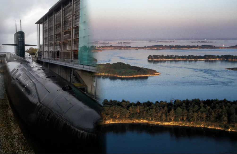 Duży czarny okręt podwodny umieszczony obok wysokiego budynku, na tle wody i wysp tworzących spokojny krajobraz, linia lasu oddziela akwen od lądu, a panorama rozciąga się aż po horyzont.