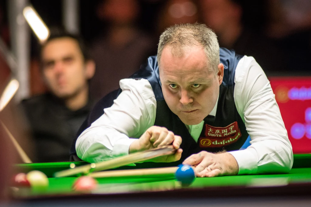 Zawodowy gracz skupiony podczas rozgrywki snookerowej, trzyma kij, celując w białą bilę na zielonym stole, w tle rozmyty obserwator.