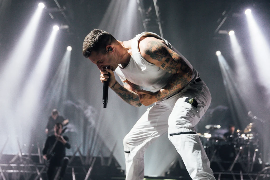 Wokalista w białym podkoszulku i spodniach z tatuażami na rękach śpiewa do mikrofonu, wyrażając silne emocje i energię podczas koncertu, w tle zespół muzyków oraz efekty świetlne sceny.