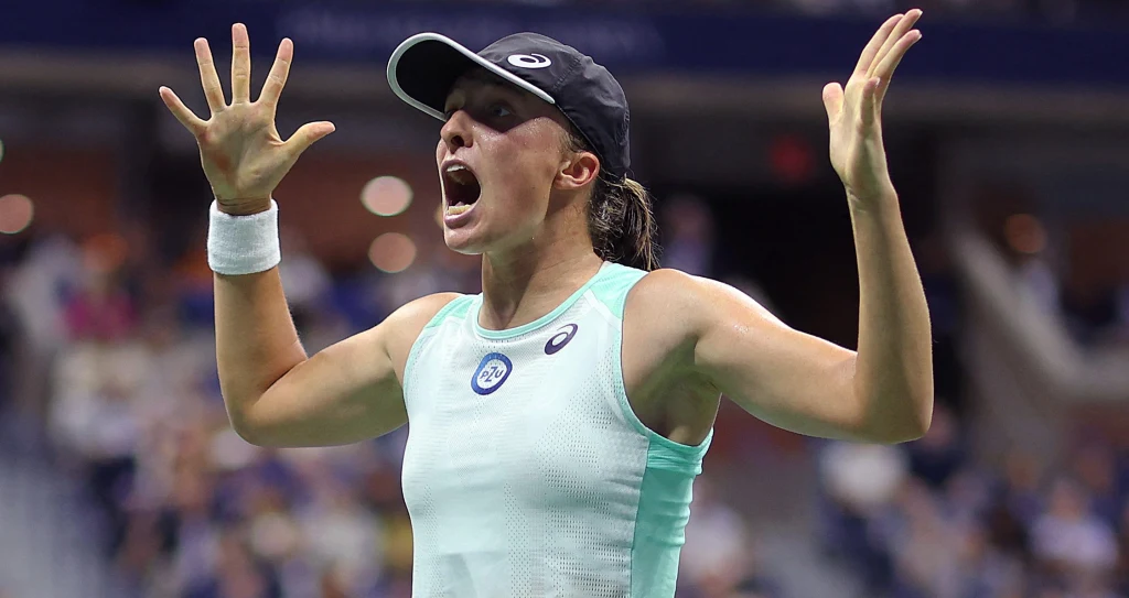 Zawodniczka tenisa w sportowej koszulce i czapce z daszkiem, w czasie intensywnej emocjonalnej reakcji, ręce uniesione w górę, szeroko otwarte usta, rozmyte tło kortu tenisowego z widoczną publicznością.
