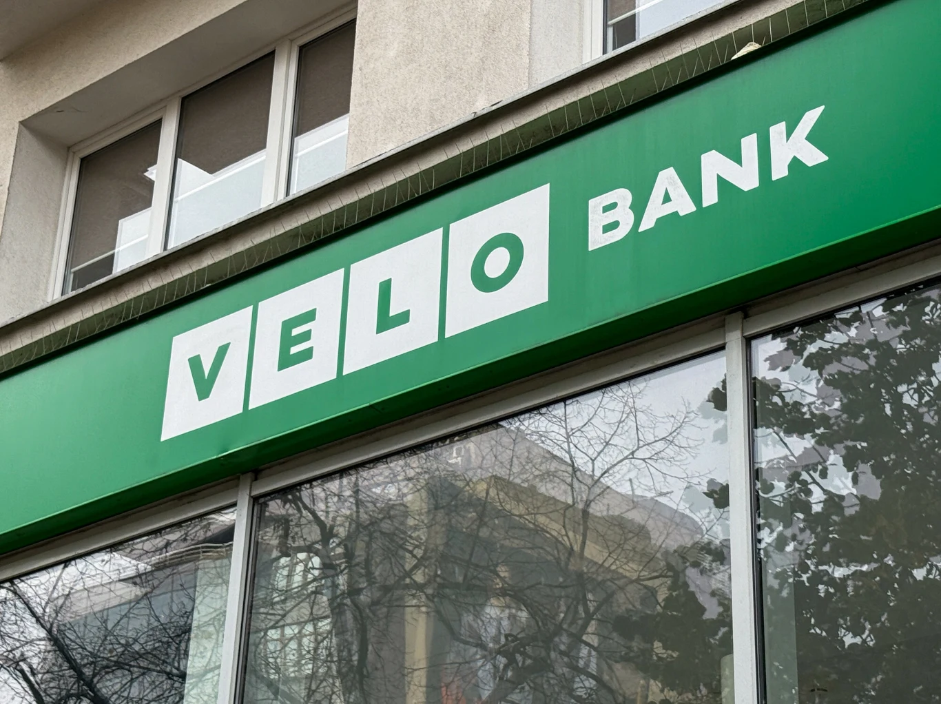 Jest zgoda KNF na przejęcie detalicznej części Citi Handlowego przez VeloBank Szyld banku Velo Bank na zielonym tle, umieszczony na szklanej fasadzie budynku z odbiciem drzew w oknach