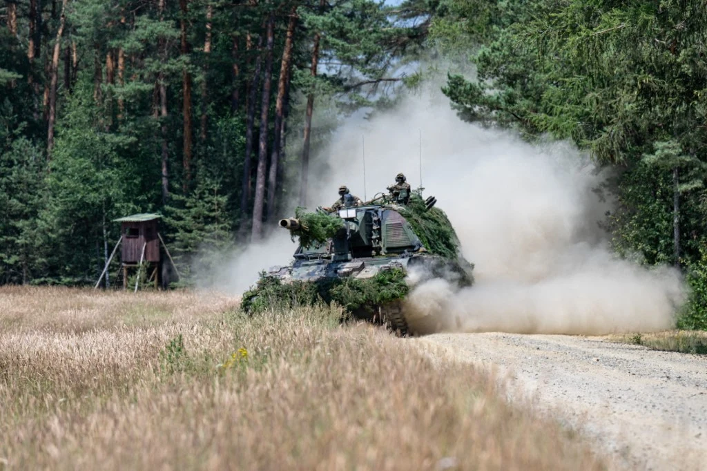 Niemiecka Bundeswehra podczas ćwiczeń Czołg porusza się z dużą prędkością polną drogą w otoczeniu wysokich traw i lasu, wzbijając za sobą gęsty obłok kurzu. W tle znajduje się drewniana ambona myśliwska wśród drzew.