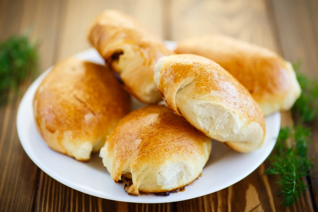 Pięć złocistych pierogów drożdżowych ułożonych na białym talerzu, na drewnianym stole, otoczonych gałązkami świeżego koperku.