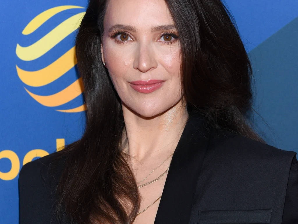 Kobieta z długimi, ciemnymi włosami, uśmiechnięta, ubrana w elegancki, czarny żakiet. W tle geometryczny wzór i żółte elementy na niebieskim tle.