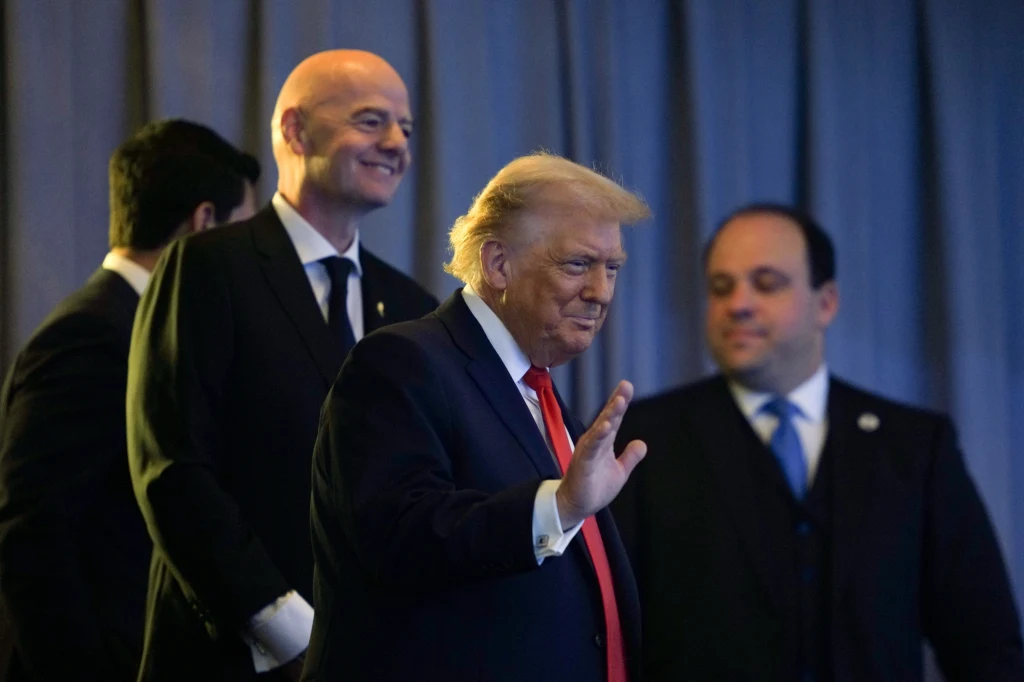 Donald Trump w towarzystwie Gianniego Infantino Mężczyzna w garniturze z pomarańczowymi włosami gestykuluje dłonią, otoczony jest przez dwóch innych mężczyzn również ubranych w garnitury. W tle widoczna jest zasłona.