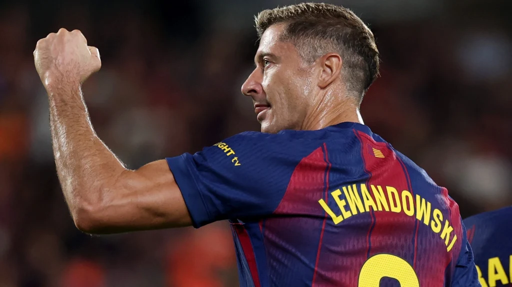 Robert Lewandowski Piłkarz w koszulce z nazwiskiem Lewandowski i numerem 9 wykonuje gest triumfu, podnosząc zaciśniętą pięść, tło rozmazane, sugerujące stadion piłkarski podczas meczu.