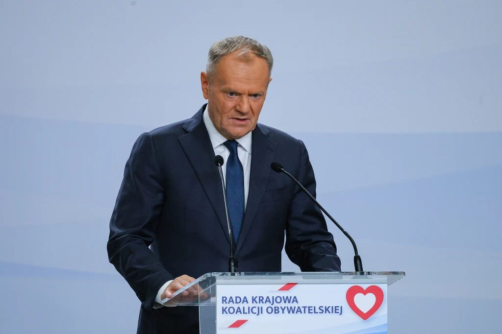 Donald Tusk Mężczyzna w garniturze wygłaszający przemówienie przy mównicy z napisem Rada Krajowa Koalicji Obywatelskiej, na tle jasnego, neutralnego tła.