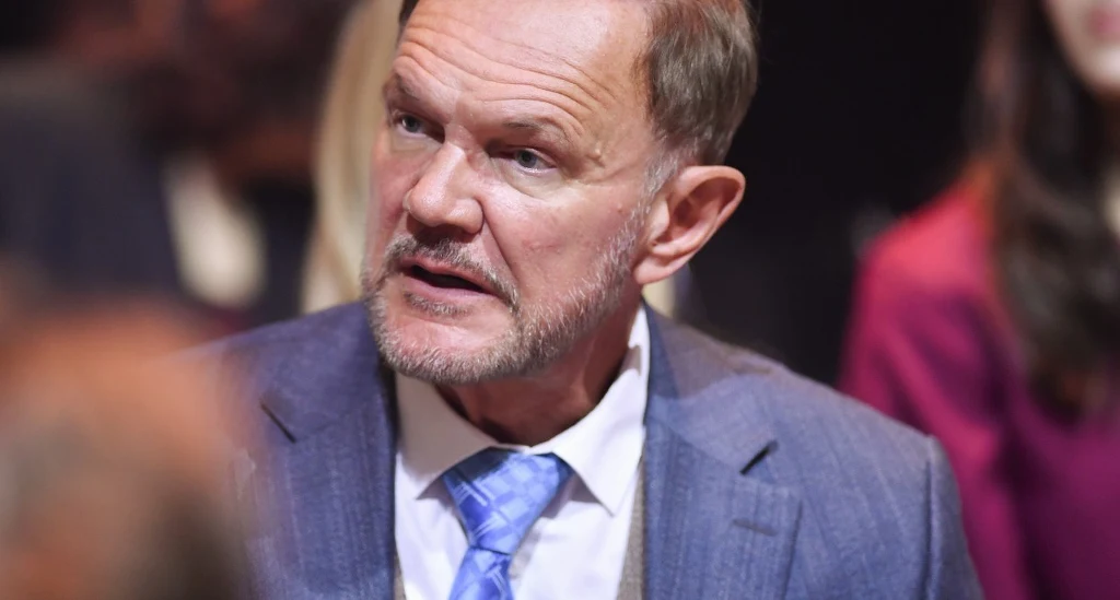 Mężczyzna w średnim wieku w garniturze i niebieskim krawacie, skierowany w bok, z poważnym wyrazem twarzy, rozmyte tło sugeruje obecność innych osób.