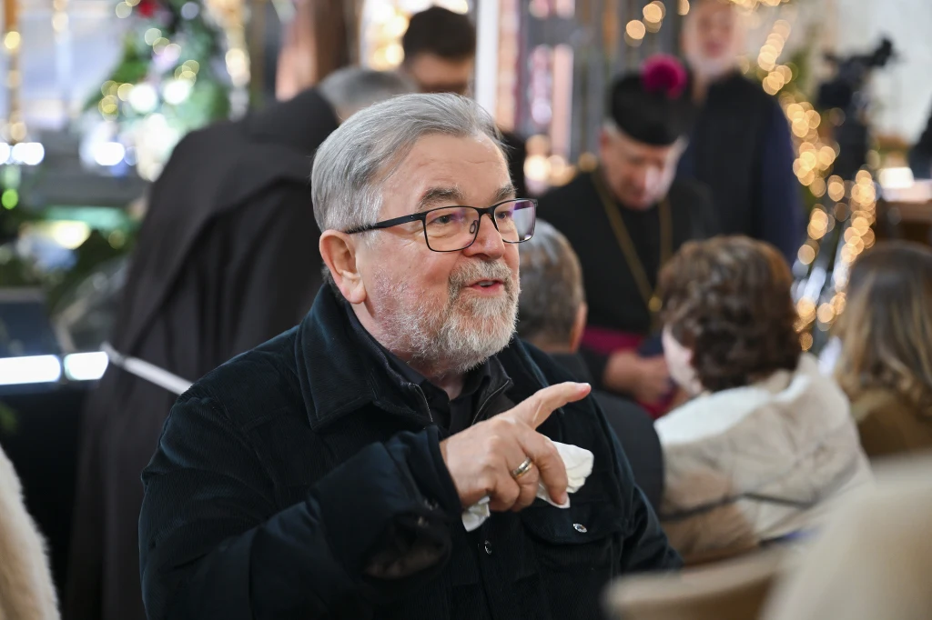 Starszy mężczyzna w okularach, z siwą brodą, rozmawia gestykulując w otoczeniu innych ludzi. W tle znajduje się kilku uczestników wydarzenia w pomieszczeniu ozdobionym światełkami, jeden z nich nosi czarny strój duchownego z charakterystyczną czarną pi...