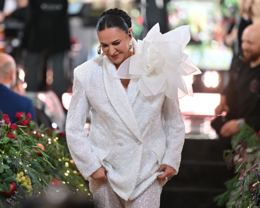 Kobieta w białym, błyszczącym garniturze ozdobionym dużym kwiatem na ramieniu, otoczona kwiatami i światłami, uśmiecha się i spogląda w dół.