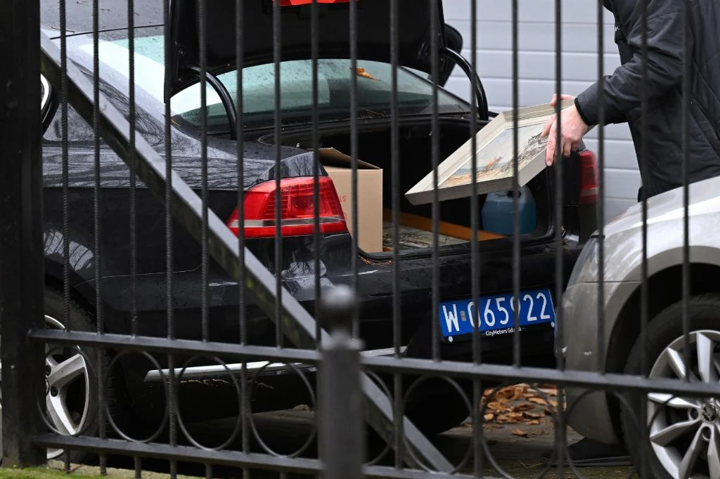 Rosjanie opuszczają konsulat w Gdańsku Mężczyzna wkłada pudełko do bagażnika czarnego samochodu zaparkowanego obok srebrnego auta, widoczna rejestracja służbowa oraz fragment metalowego ogrodzenia na pierwszym planie.