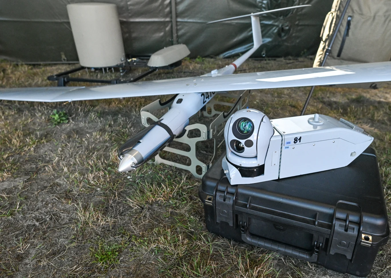 Wojskowy dron o wydłużonym skrzydle umieszczony na ziemi obok zaawansowanego modułu optycznego z kamerą, wszystko ułożone na twardej skrzynce sprzętowej na trawie w pobliżu namiotu.
