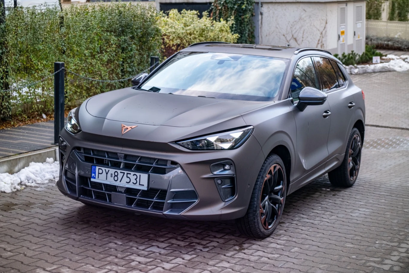 Samochód Cupra Formentor o matowym, grafitowym lakierze zaparkowany na brukowanej nawierzchni. Nowoczesny SUV charakteryzuje się wyrazistą linią nadwozia, czarnymi felgami i sportowym wyglądem. W tle ogrodzenie i zimowe pozostałości śniegu.