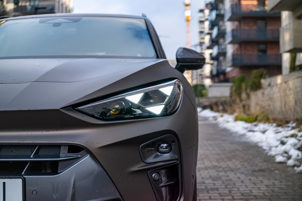 Trójkątne elementy świetlne to od jakiegoś czasu wyróżnik samochodów marki Cupra Zbliżenie na przednią część samochodu z włączonym reflektorem LED, na tle nowoczesnych budynków oraz resztek śniegu na chodniku.