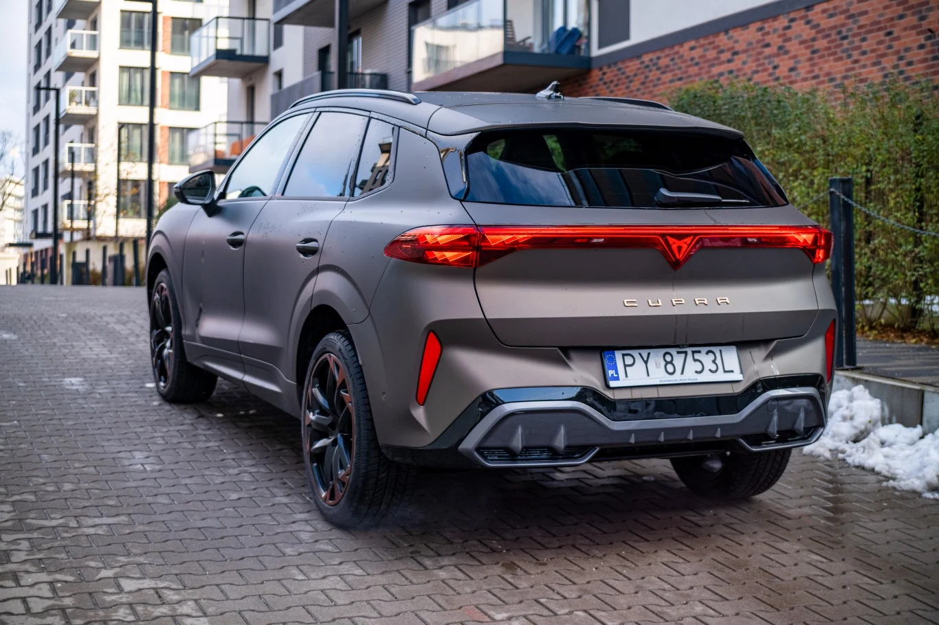 Nowoczesny SUV marki Cupra o matowym lakierze, zaparkowany na brukowanej ulicy wśród nowoczesnych budynków mieszkalnych, widok z tyłu ukazujący charakterystyczne tylne światła oraz polską tablicę rejestracyjną.