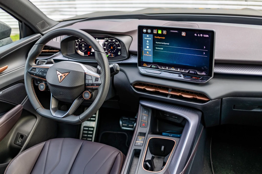 Wnętrze jest sensownie zaprojektowane, a kokpit – łatwy w obsłudze Kokpit nowoczesnego samochodu marki Cupra, z cyfrowym zestawem wskaźników, dużym ekranem dotykowym systemu multimedialnego oraz sportową kierownicą z charakterystycznym logo producenta. Wnętrze wyposażone w podświetlane przyciski, wysokiej jakości wyko...