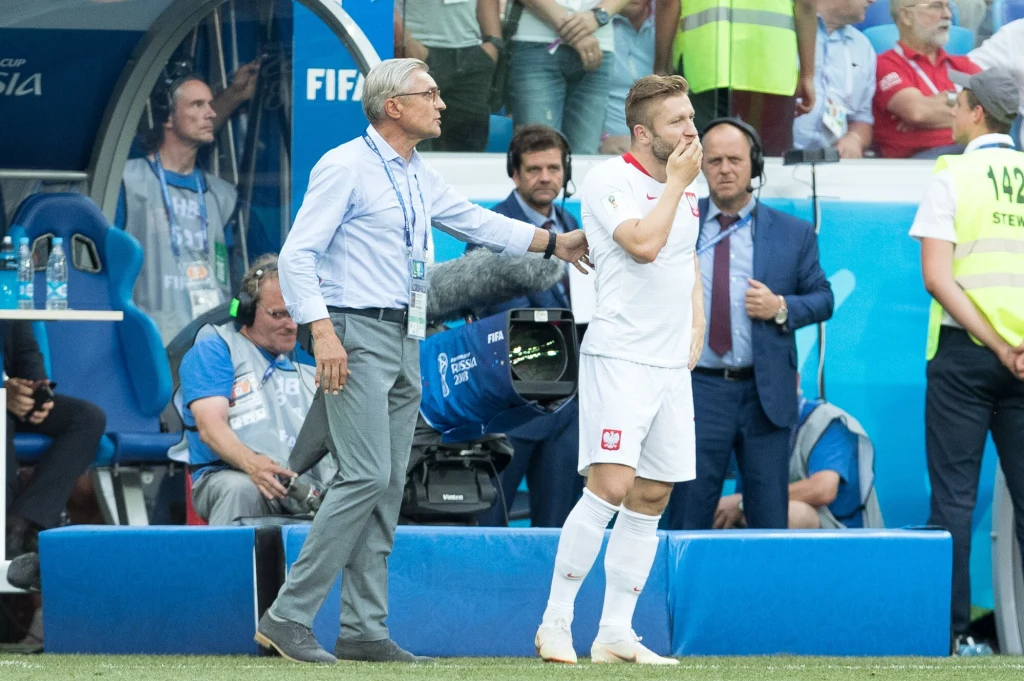 Polska znów ma szansę zagrać z Japonią Trener dotyka ramienia piłkarza reprezentacji Polski na linii bocznej podczas meczu, piłkarz wygląda na zmartwionego lub rozczarowanego. W tle widać innych członków sztabu oraz kibiców.