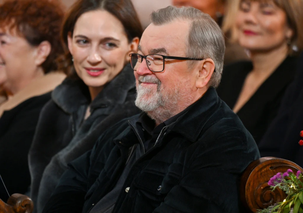 Starszy mężczyzna z siwą brodą i okularami siedzi uśmiechnięty wśród innych ludzi, za nim znajduje się młoda kobieta z przyjaznym wyrazem twarzy, w tle widoczne są rozmyte postaci innych uczestników spotkania.