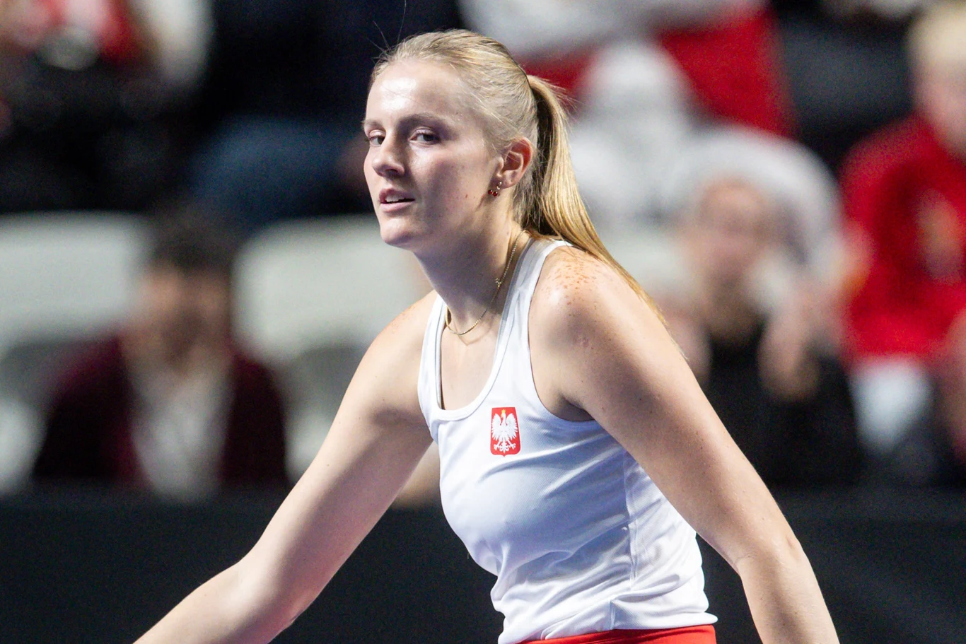 Kobieta w sportowym stroju z biało-czerwoną naszywką z godłem Polski na tle kibiców na trybunach. Na jej twarzy widoczny jest wysiłek po intensywnej aktywności fizycznej.