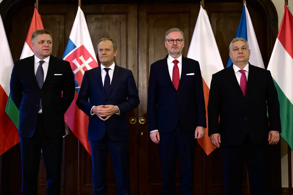Czterech mężczyzn ubranych w garnitury stoi na tle flag Polski, Słowacji, Czech i Węgier, ustawionych na drewnianym tle, oficjalna atmosfera spotkania politycznego.