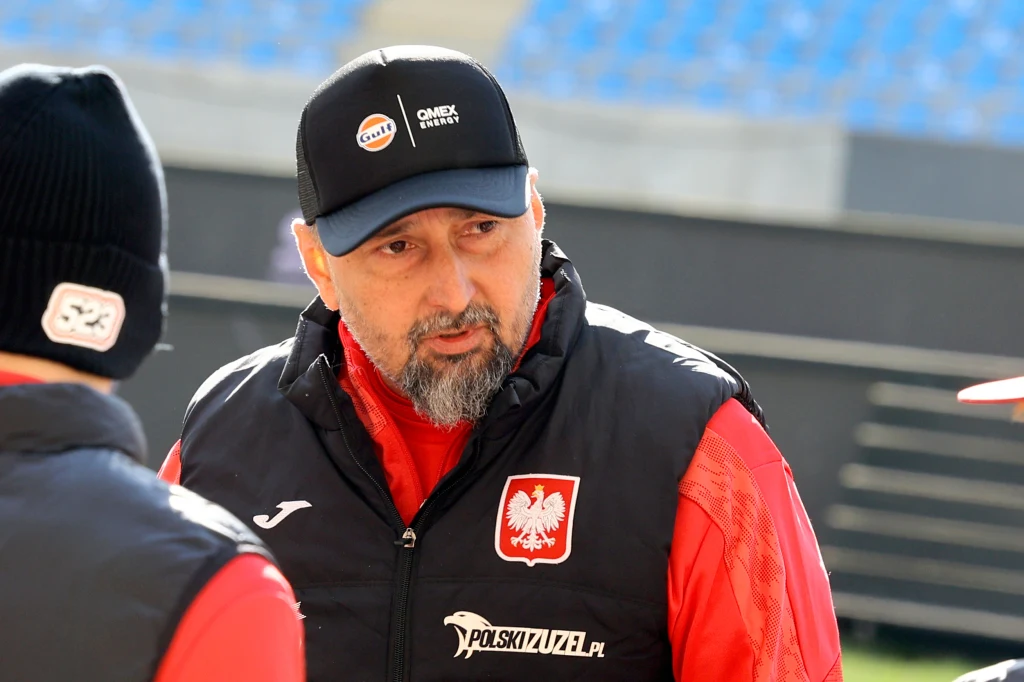 Rafał Dobrucki Mężczyzna z brodą w czapce z daszkiem i kurtce z godłem Polski rozmawia z innymi osobami ubranego w sportowe stroje na stadionie.