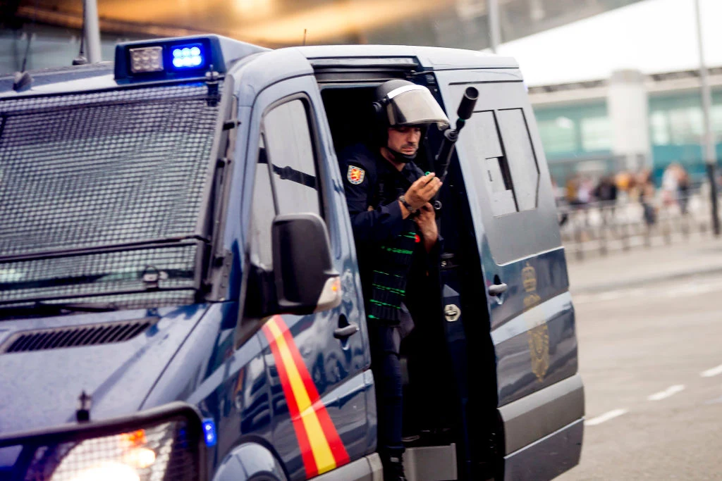 Hiszpańska policja zatrzymała Polaka. Zarzuty o udział w zorganizowanej grupie przestępczej Policjant w stroju ochronnym stoi w otwartych drzwiach policyjnego samochodu, trzymając broń, na tle ulicy i miejskiej infrastruktury.