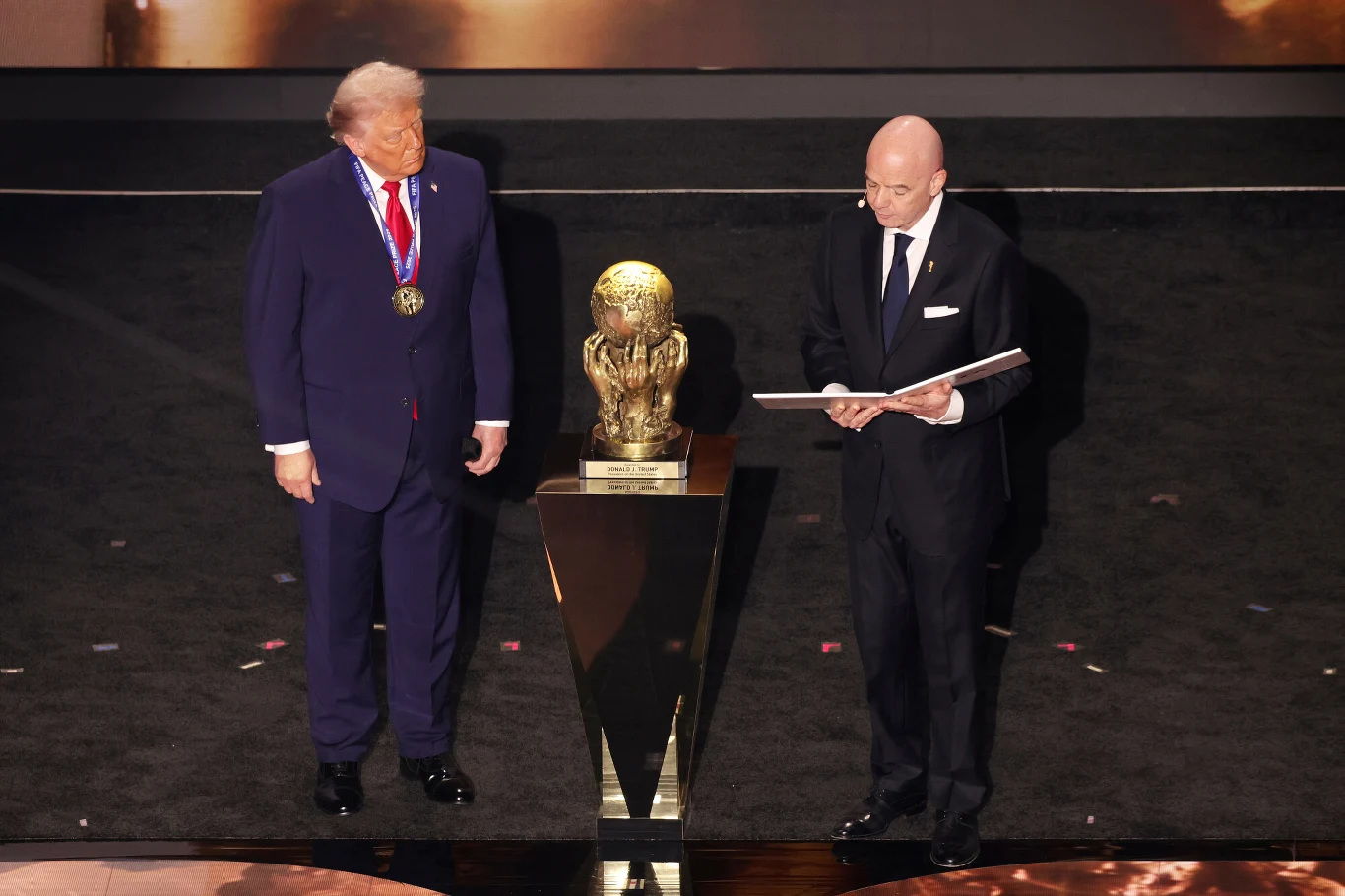 Dwóch elegancko ubranych mężczyzn, jeden z nich z medalem na szyi, stoi na scenie obok dużego trofeum w kształcie piłki na postumencie, drugi trzyma otwartą księgę i czyta.