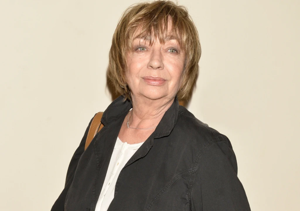 Kobieta o krótkich, jasnobrązowych włosach, ubrana w ciemną marynarkę oraz białą bluzkę, patrzy w stronę obiektywu z delikatnym uśmiechem; tło jednolite, jasne.