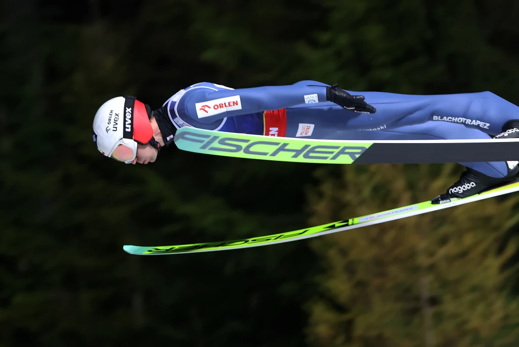 Sportowiec w kombinezonie do skoków narciarskich podczas lotu w powietrzu, trzymający sylwetkę aerodynamiczną, jasne narty Fischer, w tle rozmyte drzewa.