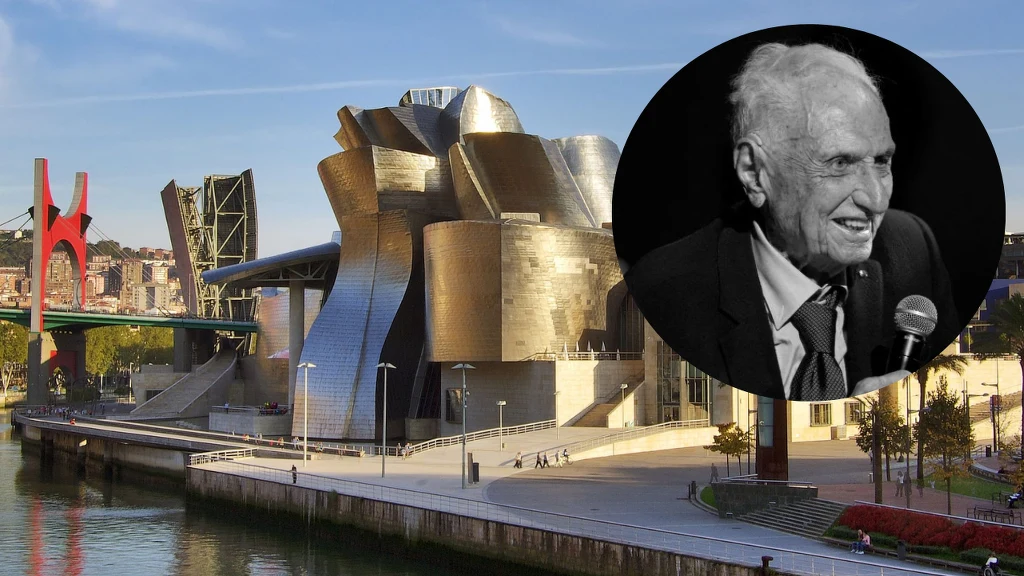 Nowoczesny budynek o nieregularnych, metalicznych kształtach odbijających światło słoneczne nad rzeką, w tle most oraz fragmenty miasta. W prawym górnym rogu czarno-białe zdjęcie starszego mężczyzny uśmiechającego się do mikrofonu.