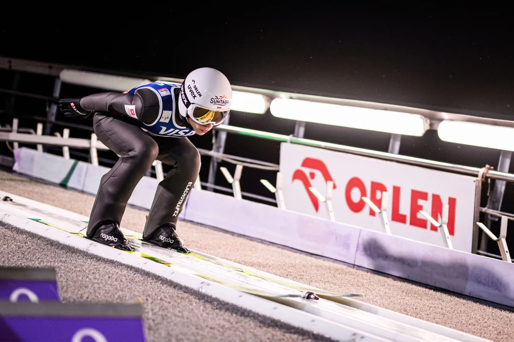 Aleksander Zniszczoł Skoczek narciarski w pozycji najazdowej na rozbiegu, ubrany w kombinezon sportowy i kask, na tle oświetlonej skoczni i reklamy Orlen.