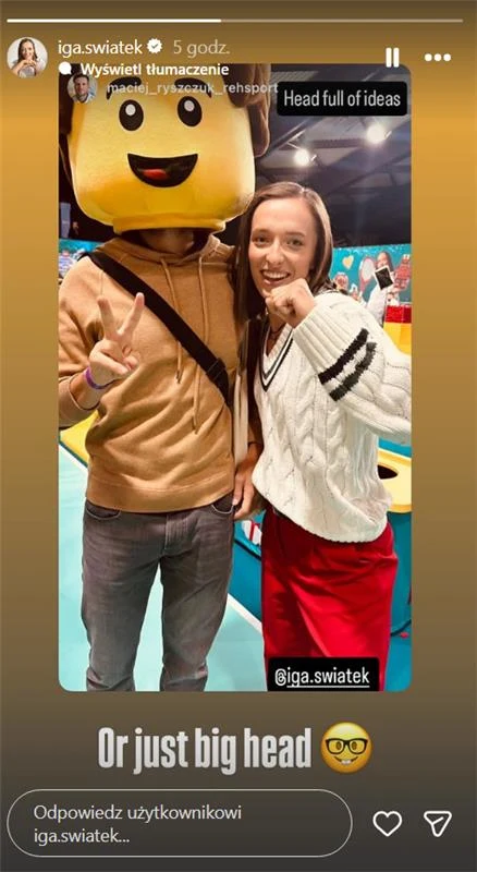 Dwie osoby pozujące do zdjęcia, jedna z nich ma dużą maskę w kształcie klocka LEGO na głowie, obok niej stoi kobieta uśmiechająca się do aparatu, w tle kolorowe elementy dekoracyjne i tłum ludzi.