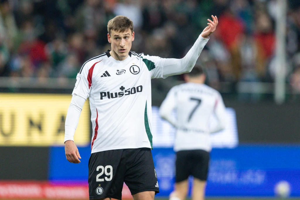 Mileta Rajović Piłkarz w białej koszulce z numerem 29 na piersi stoi na boisku, podnosi rękę w geście komunikacji podczas meczu. W tle widoczny inny zawodnik w podobnej koszulce oraz rozmazana publiczność.