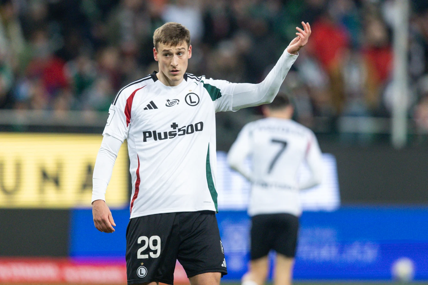 Mileta Rajović Piłkarz w białej koszulce z numerem 29 na piersi stoi na boisku, podnosi rękę w geście komunikacji podczas meczu. W tle widoczny inny zawodnik w podobnej koszulce oraz rozmazana publiczność.