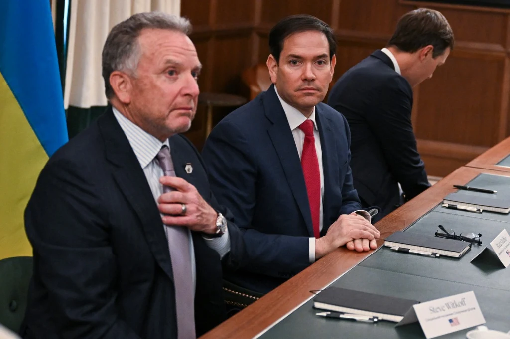 Dwaj mężczyźni w garniturach siedzą przy stole konferencyjnym z dokumentami, tabliczkami z nazwiskami i mikrofonami, obok nich widoczny fragment flagi Ukrainy.