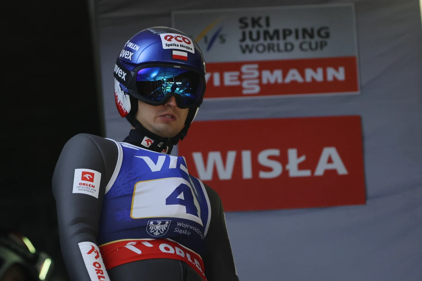 Maciej Kot Zawodnik w kombinezonie narciarskim i kasku z goglami, przygotowany do skoku narciarskiego na tle tablicy informującej o zawodach Pucharu Świata w Wiśle.