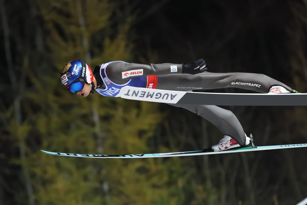 Skoczek narciarski w locie podczas zawodów na tle rozmytego, ciemnego lasu. Zawodnik ma na sobie profesjonalny kombinezon, kask oraz gogle ochronne. Narty są wyciągnięte równolegle do ziemi, a całość ukazuje moment maksymalnego skupienia i dynamiki.