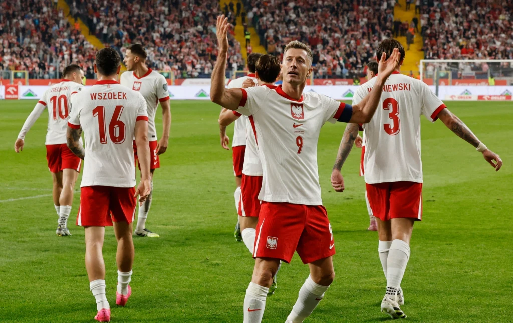 Piłkarze reprezentacji Polski w białych koszulkach i czerwonych spodenkach świętują na boisku, wokół nich tłum kibiców na trybunach. Na pierwszym planie zawodnik z numerem 9 z uniesionymi ramionami, wokół inni gracze drużyny.