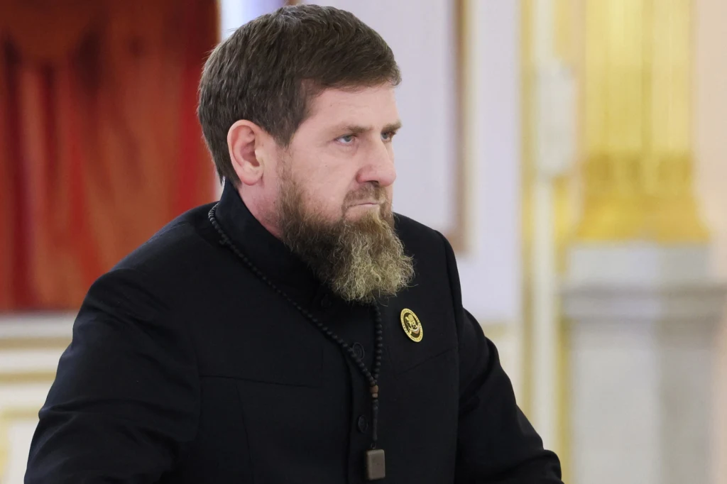 Mężczyzna w średnim wieku z brodą, ubrany w ciemny garnitur ze złotą przypinką, stoi w eleganckim, jasnym wnętrzu z ozdobnymi złotymi elementami.