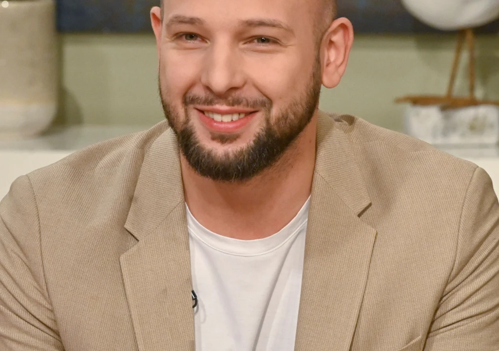 Maciej Pela Uśmiechnięty mężczyzna z brodą i krótkimi włosami ubrany w jasny beżowy marynarkę oraz biały t-shirt, siedzi przy stole w jasnym, nowoczesnym wnętrzu.