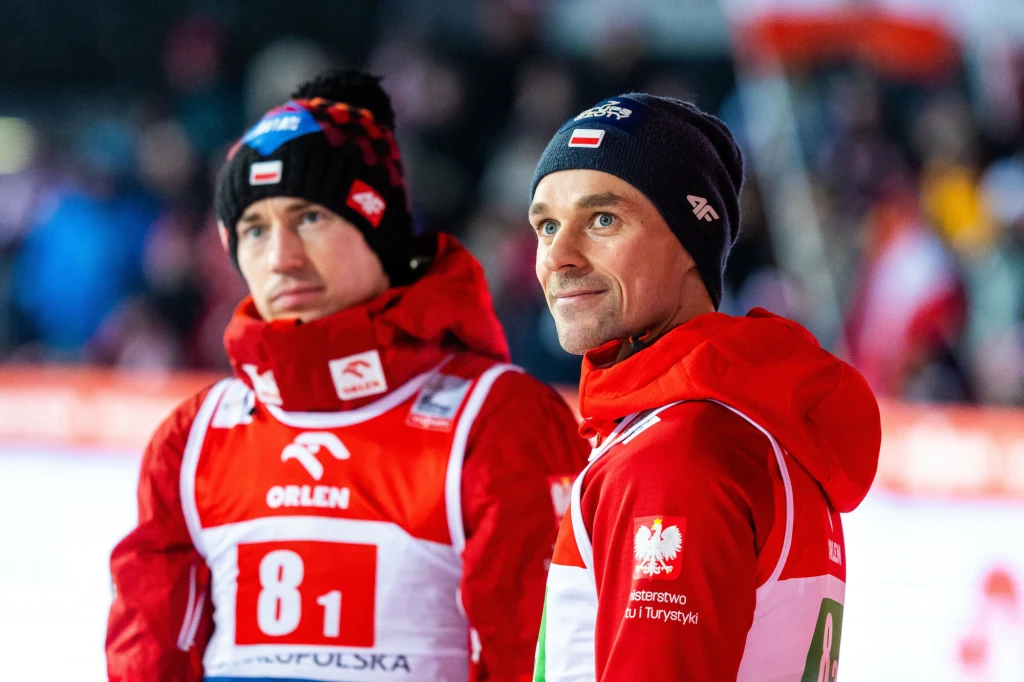 Kamil Stoch i Piotr Żyła Dwóch polskich skoczków narciarskich w czerwonych kurtkach i czapkach z biało-czerwonymi emblematami, stojących na tle rozświetlonego stadionu, z widocznymi numerami startowymi oraz logo sponsorów.