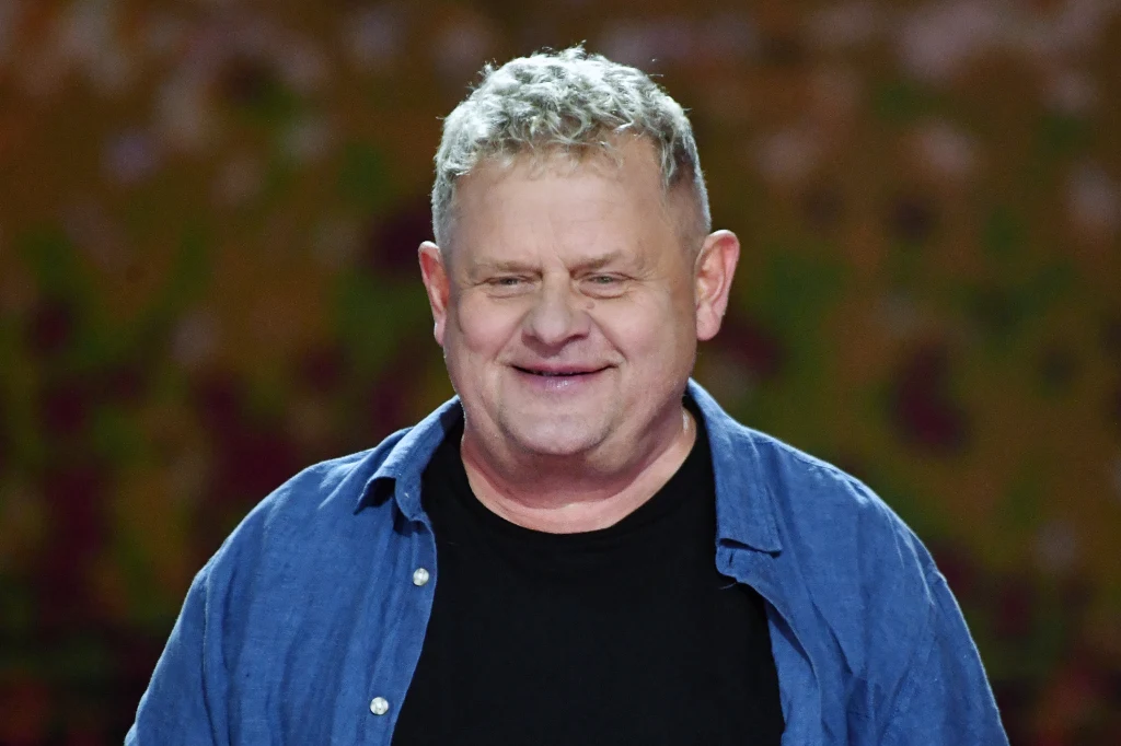 Kazik wystosował wiadomość do fanów Mężczyzna w średnim wieku z krótkimi, siwymi włosami oraz uśmiechem na twarzy, ubrany w niebieską koszulę i ciemny t-shirt, na rozmytym tle w ciepłych kolorach.