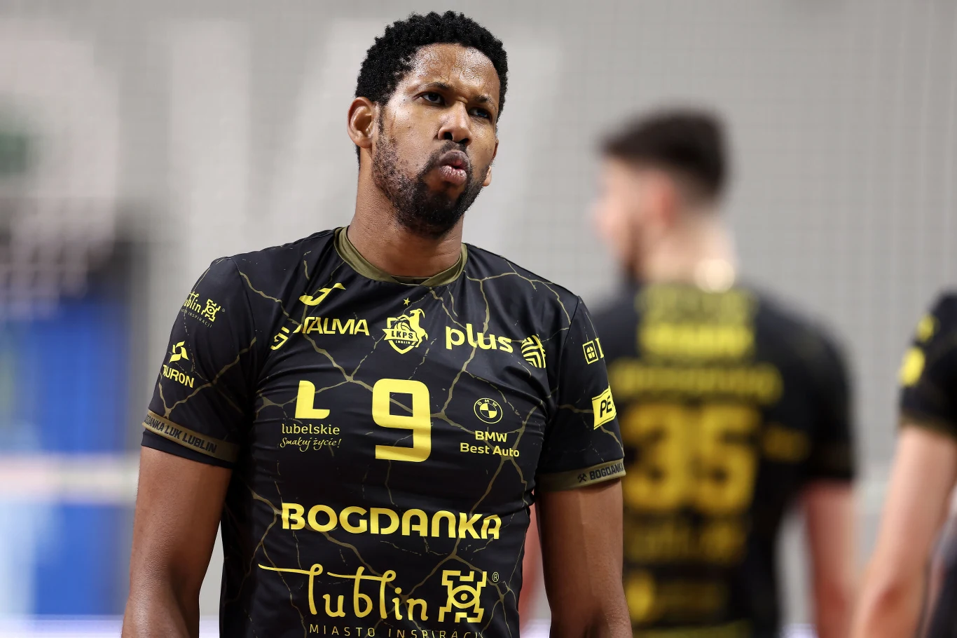 Siatkarz drużyny sportowej w jasnozielonym stroju z numerem 18 dynamicznie reaguje podczas rozgrywki, otoczony innymi graczami na tle boiska sportowego.