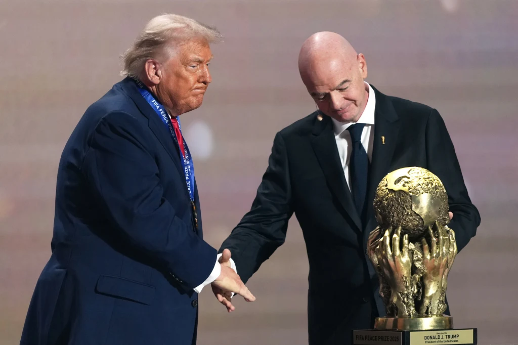 Donald Trump i Gianni Infantino, 05.12.2025 r. Dwóch mężczyzn w garniturach wymienia uścisk dłoni, obok nich znajduje się duża złota statuetka przypominająca puchar świata, jeden z mężczyzn patrzy w dół, drugi utrzymuje kontakt wzrokowy z rozmówcą.