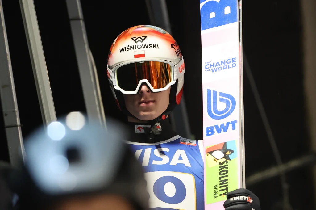 Skoczek narciarski w kombinezonie sportowym, hełmie i goglach przygotowujący się do oddania skoku narciarskiego. Na tle widoczne elementy skoczni i reklamy sponsorów.