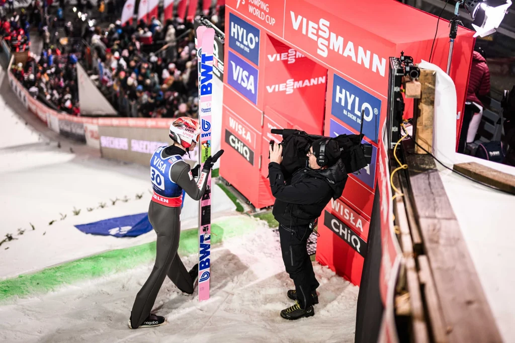 Zawodnik skoków narciarskich w kombinezonie oraz kasku trzyma narty i udziela wywiadu kamerzyście po zakończonym skoku, w tle widoczna jest trybuna z kibicami oraz reklamy sponsorskie.